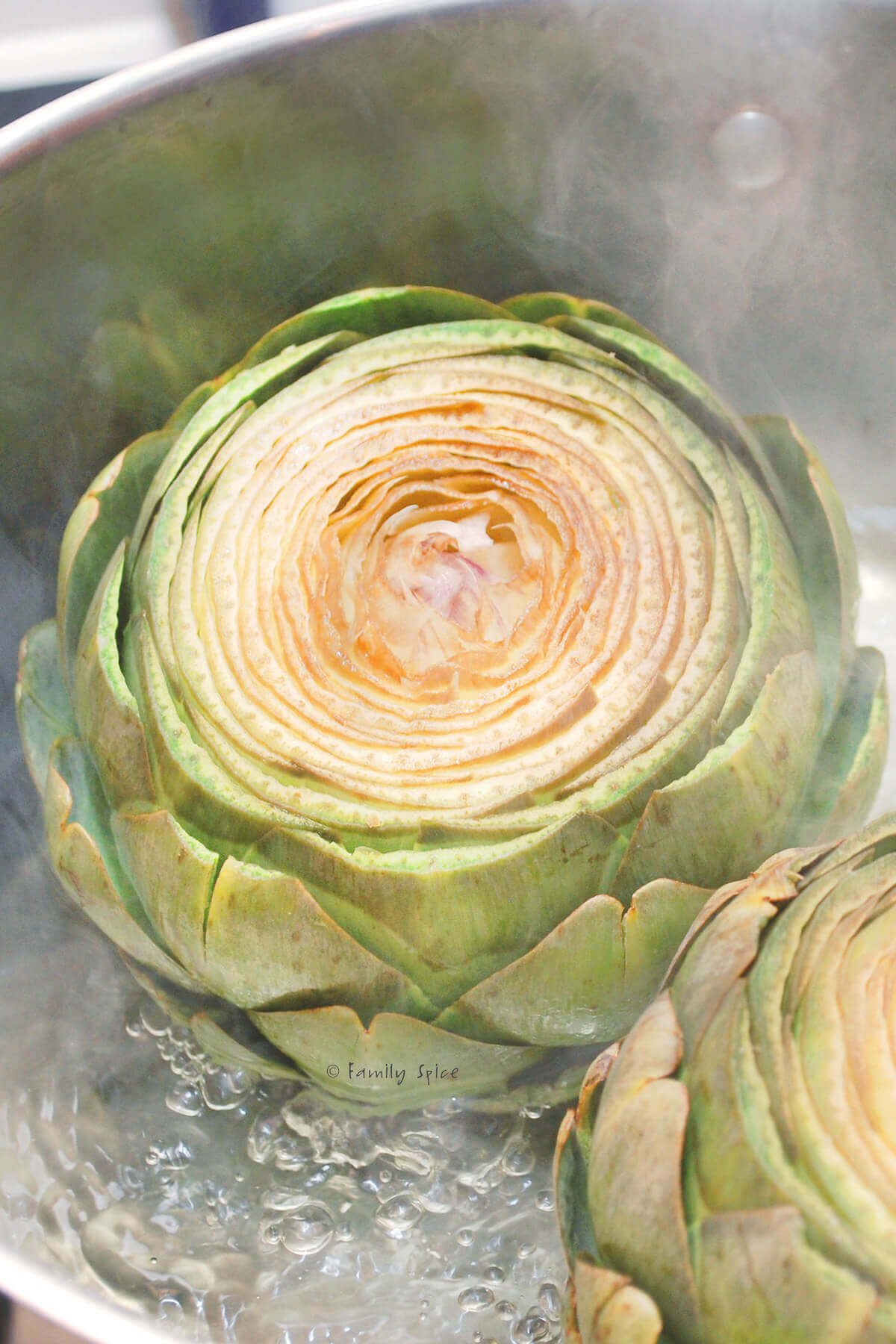 An artichoke with the top cut off steaming in a pot with hot water
