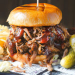 A stuffed pulled pork sandwich dripping with barbecue sauce and served with chips and pickles by FamilySpice.com