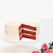 A beet red velvet cake on a white cake stand with pink roses next to it