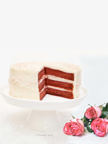 A beet red velvet cake on a white cake stand with pink roses next to it