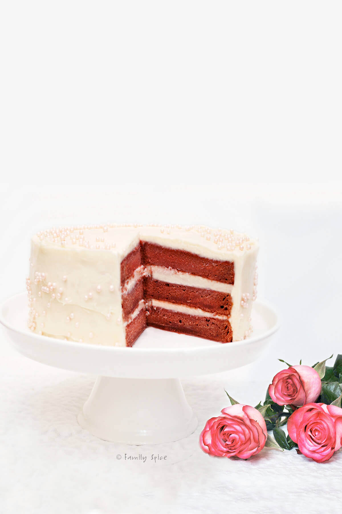 A beet red velvet cake on a white cake stand with pink roses next to it
