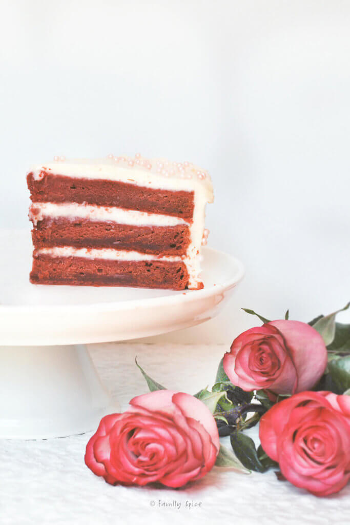 A slice of beet red velvet cake on a white cake stand with pink roses next to it
