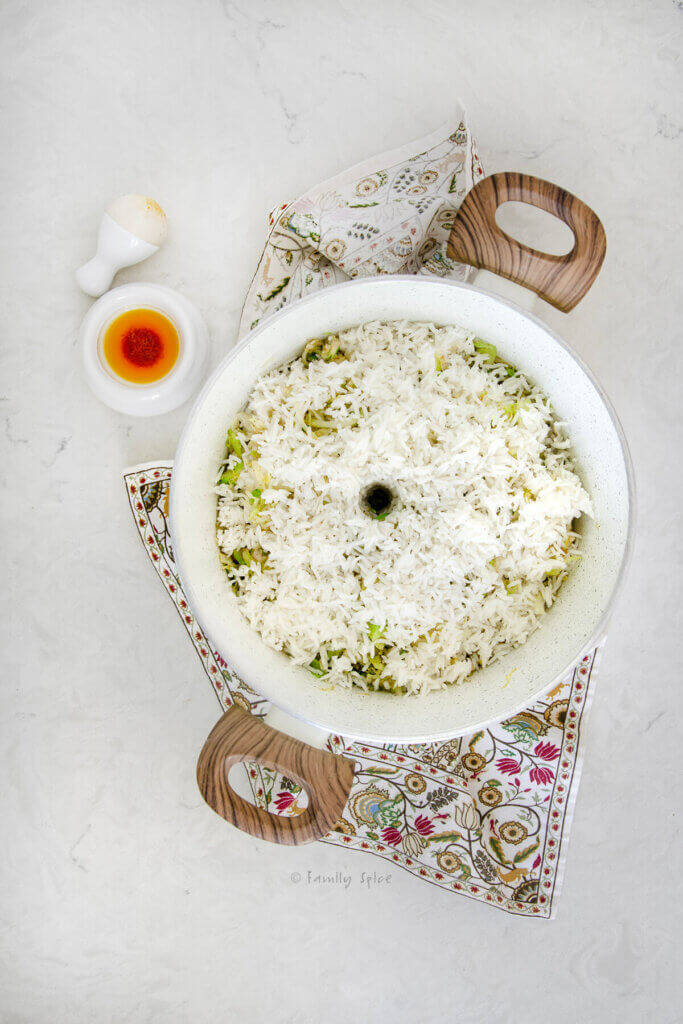 Top view of a small pot filled with parboiled basmati rice and sautéed cabbage layered in it, a hole poked down the center and with a mortal and pestle with saffron liquid spoon next to it