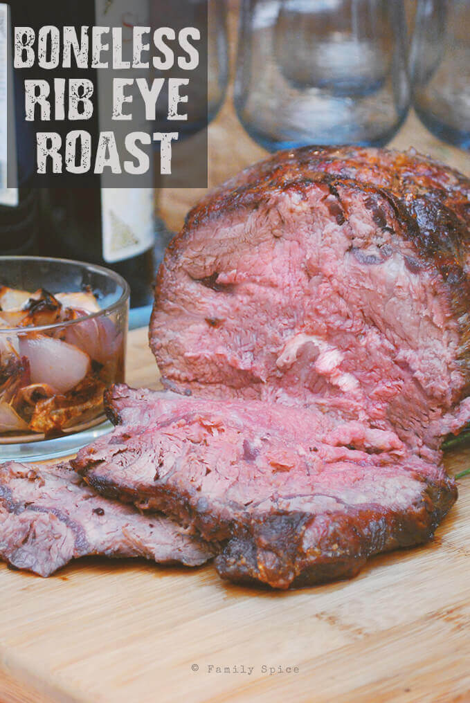 Boneless rib eye roast on a wooden cutting board with a slice cut from it and a bowl of caramelized shallots