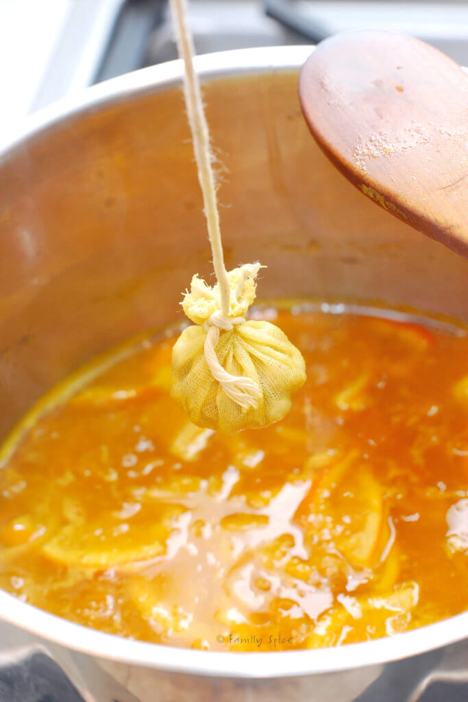 A small sack made with cheese cloth holding orange seeds and held over a pot of bubbling orange marmalade