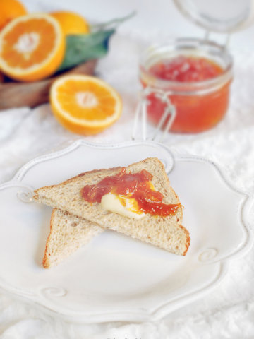 Orange jam spread on toast with a jar of orange marmalade and oranges behind it