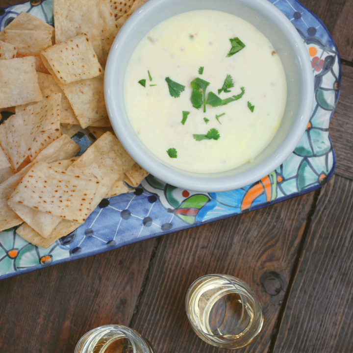 Top view of a bowl of queso in a decorated rectangular platter with tortilla chips and shot glasses with tequila