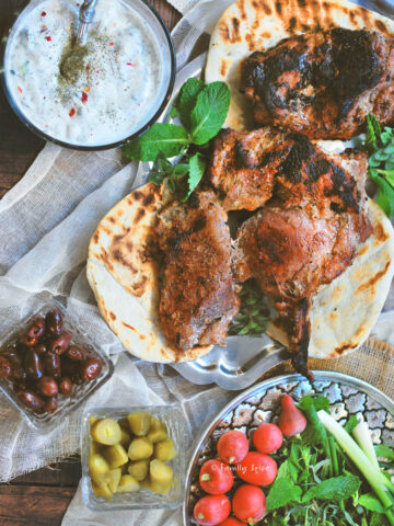 Top view of a table with grilled boneless leg of lamb on a platte with fresh herbs, radishes, flat bread, yogurt, olives and pickles