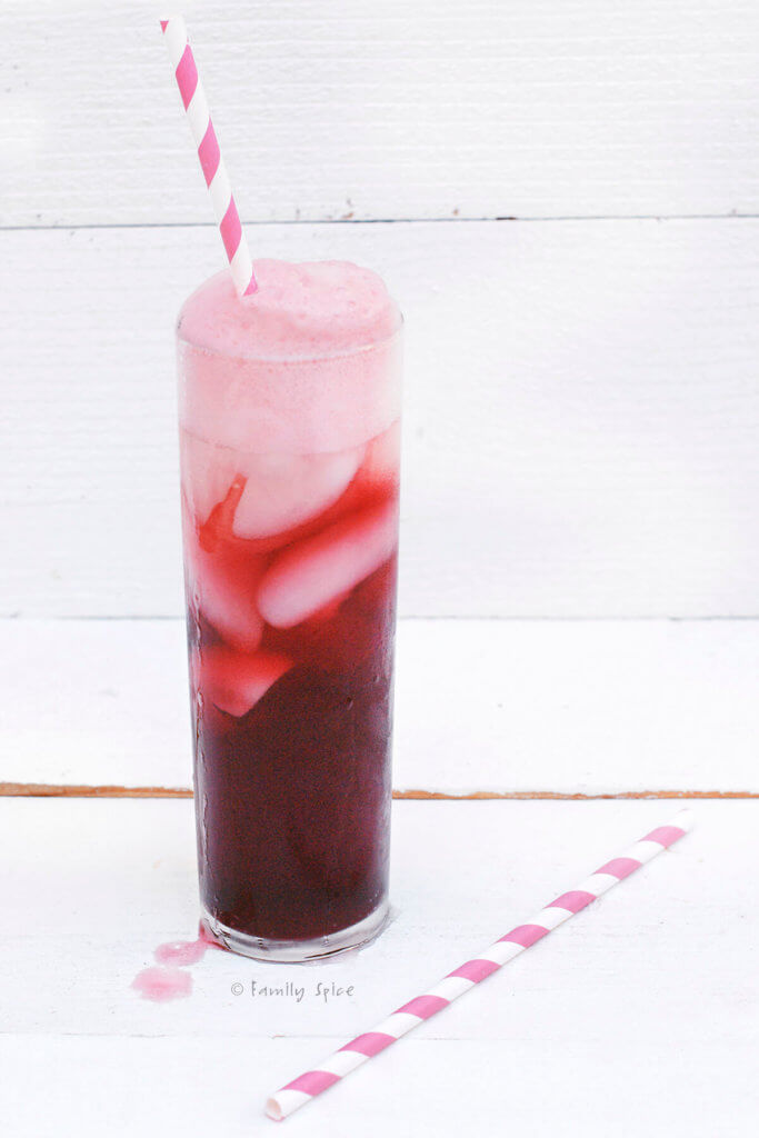 A tall glass of foamy pomegranate shirley temple with striped straw on a white background