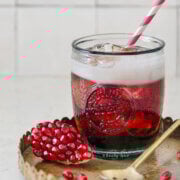 A glass with a pomegranate Shirley Temple drink and straw, with pomegranate arils next to it.