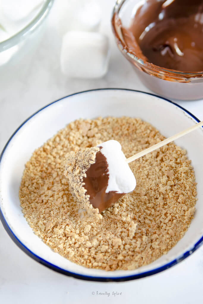 Coating a chocolate dipped marshmallow on a stick into a plate of graham cracker crumbs