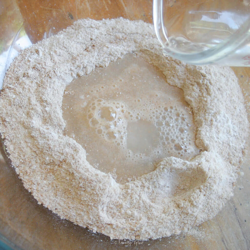 Whole wheat flour in a glass bowl with water yeast mixture being poured in