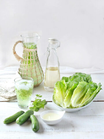 A pitcher of sekanjabeen with a bottle of syrup, a platter of romaine lettuce, cucumbers and a glass of mint cooler