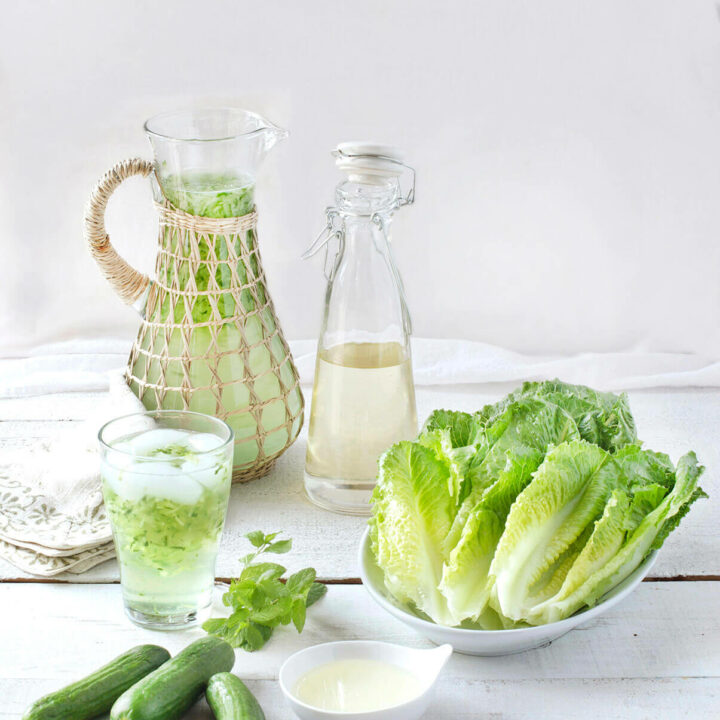 A pitcher of sekanjabeen with a bottle of syrup, a platter of romaine lettuce, cucumbers and a glass of mint cooler