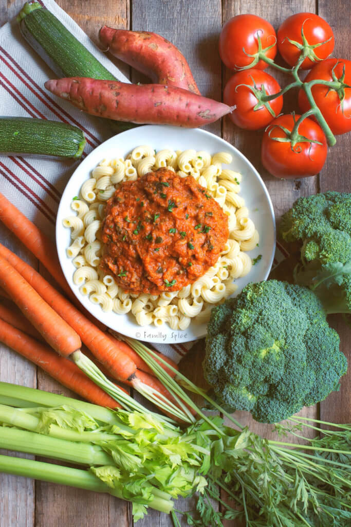 A bowl of pasta with red sauce over it surrounded by various fresh vegetables