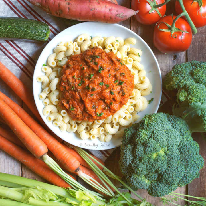 A bowl of pasta with red sauce over it surrounded by various fresh vegetables