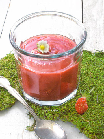 A glass jar with strawberry apple sauce topped with a strawberry flower on a bed of moss