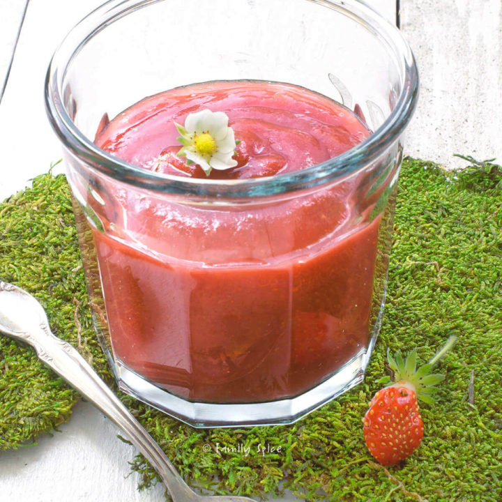 A glass jar with strawberry apple sauce topped with a strawberry flower on a bed of moss