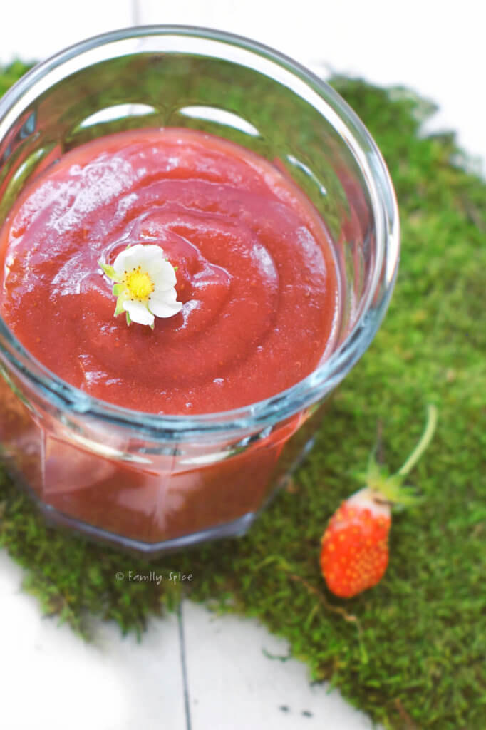 Top view of a glass jar with strawberry apple sauce topped with a strawberry flower on a bed of moss