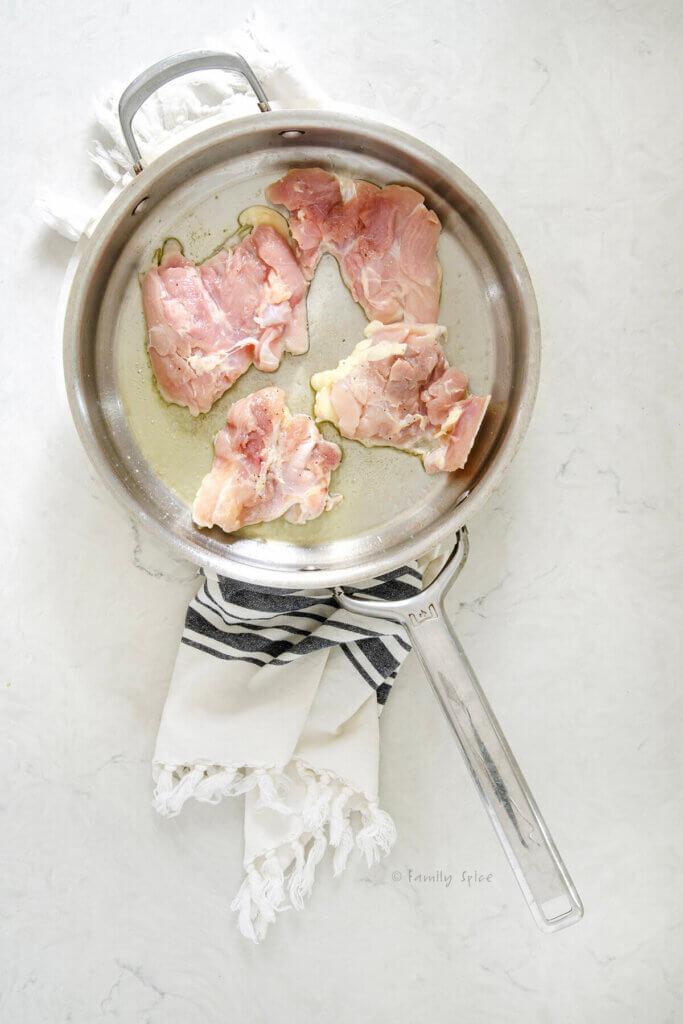 A stainless pan with olive oil and four raw boneless skinless chicken thighs in it