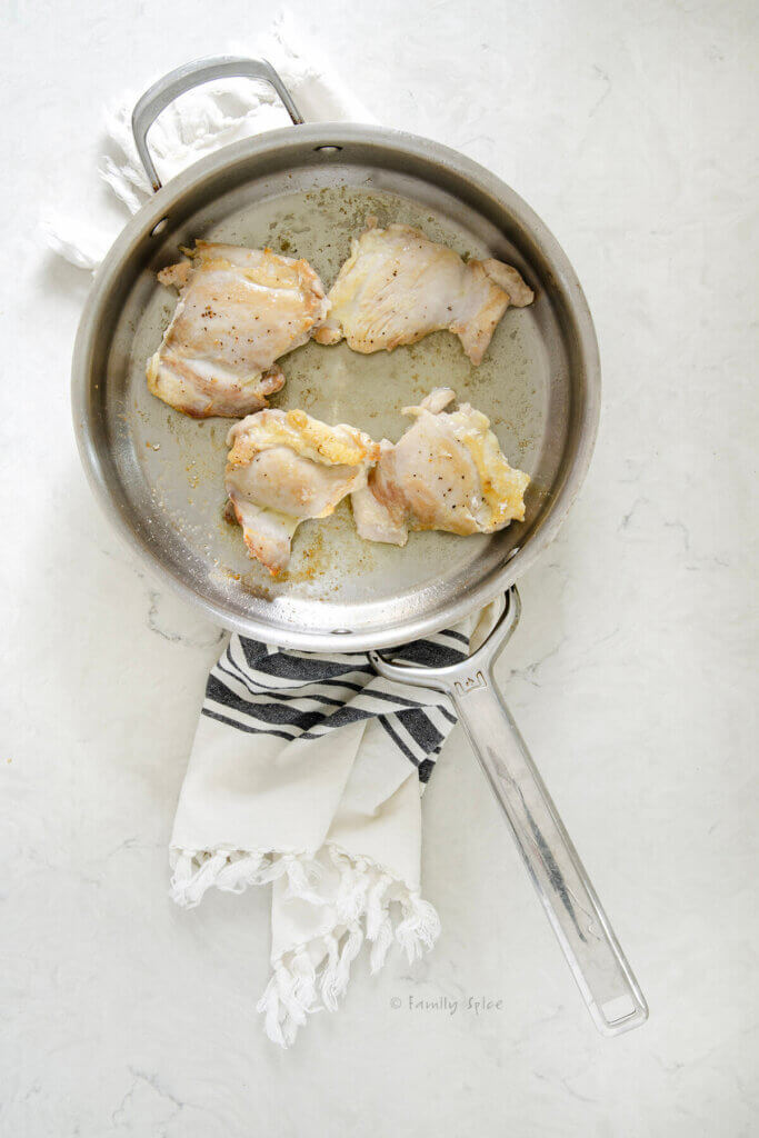 A stainless pan with olive oil and four browned boneless skinless chicken thighs in it