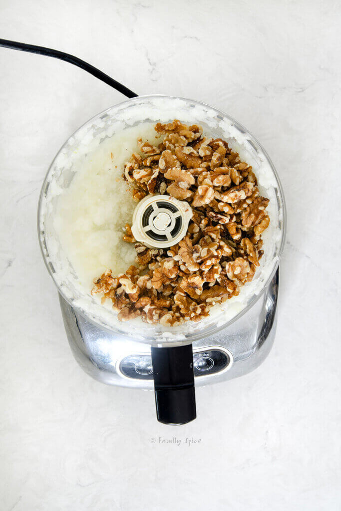 Top view of a food processor bowl with processed onions and whole walnuts in it