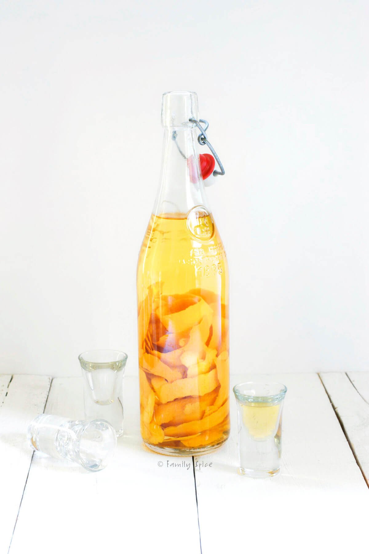 A bottle of orange vodka with orange peels in it and shot glasses next to it