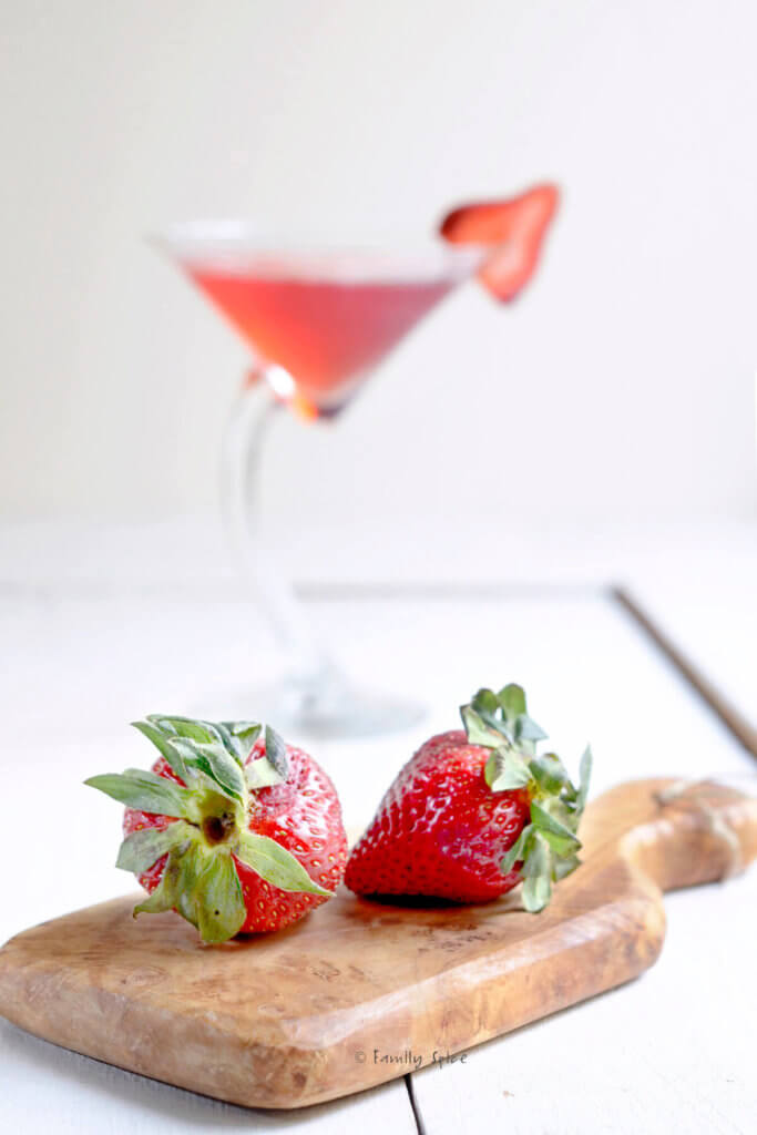 Closeup of a small wooden cutting board with two strawberries on it and a crooked martini glass with strawberry martini in it behind it