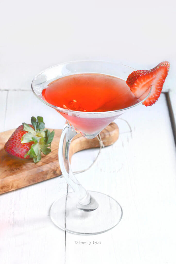 ¾ view of a crooked martini glass with strawberry martini in it and a slice of strawberry on the rim