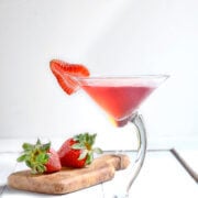 Side view of a crooked martini glass with strawberry martini in it and a slice of strawberry on the rim and strawberries behind it