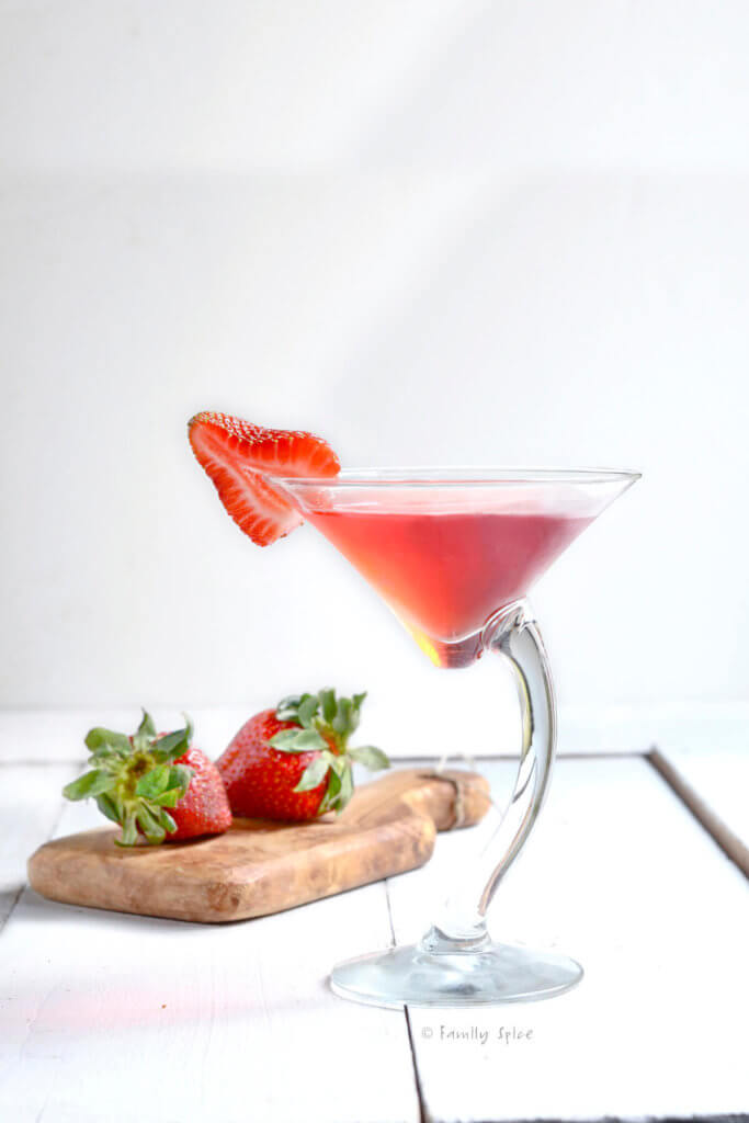 Side view of a crooked martini glass with strawberry martini in it and a slice of strawberry on the rim and strawberries behind it