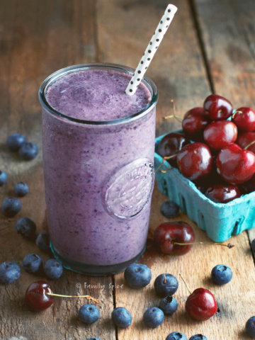 A tall glass of purple smoothie with cherries and blueberries around it