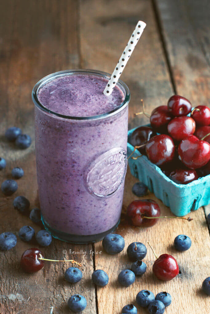 A tall glass of purple smoothie with cherries and blueberries around it