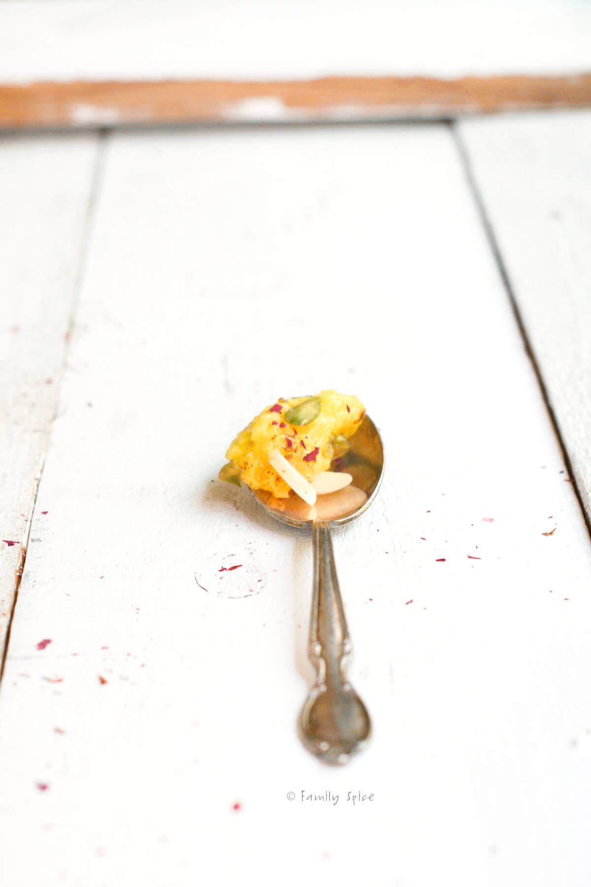 An ornate silver spoon holding shole zard garnished with cinnamon, rose petals, pistachios and almonds