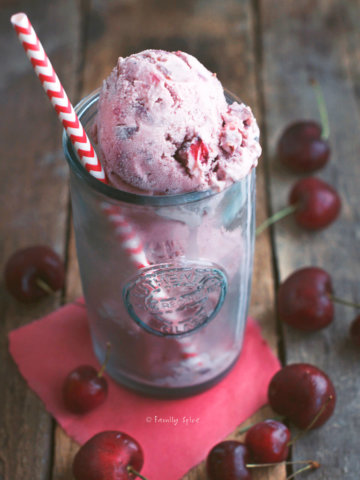 Three scoops of cherry cola ice cream in a tall glass with a straw and cherries around it