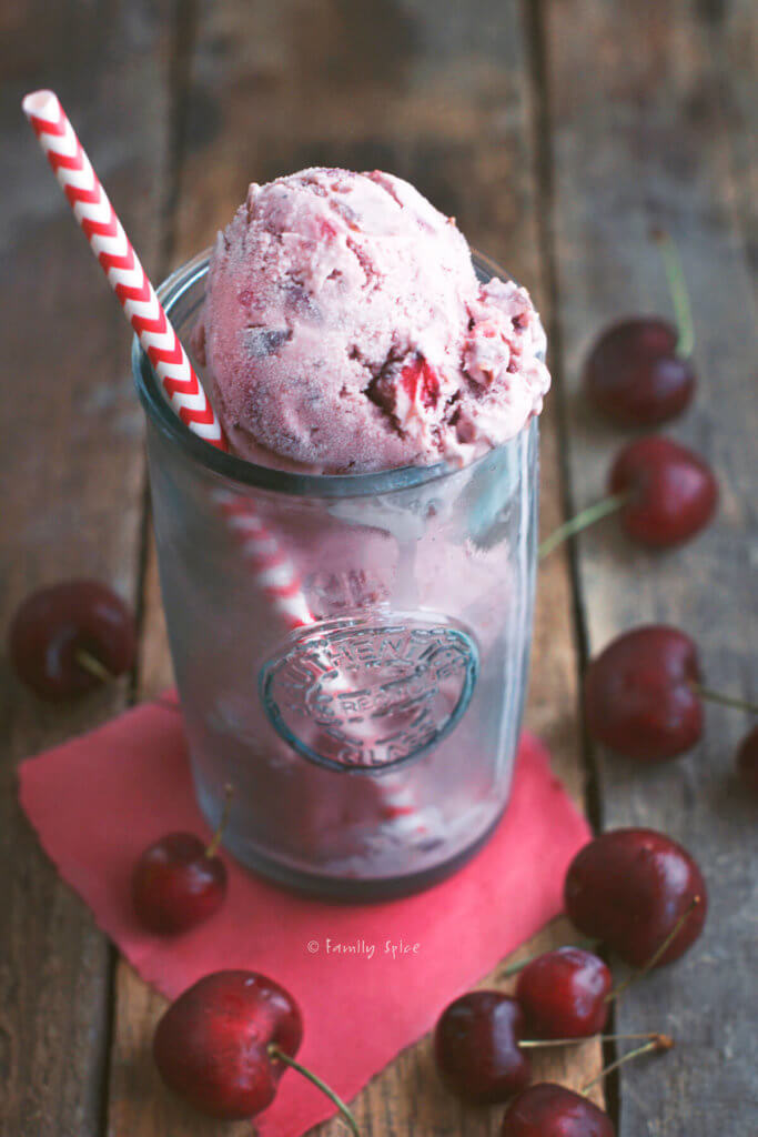 Three scoops of cherry cola ice cream in a tall glass with a straw and cherries around it