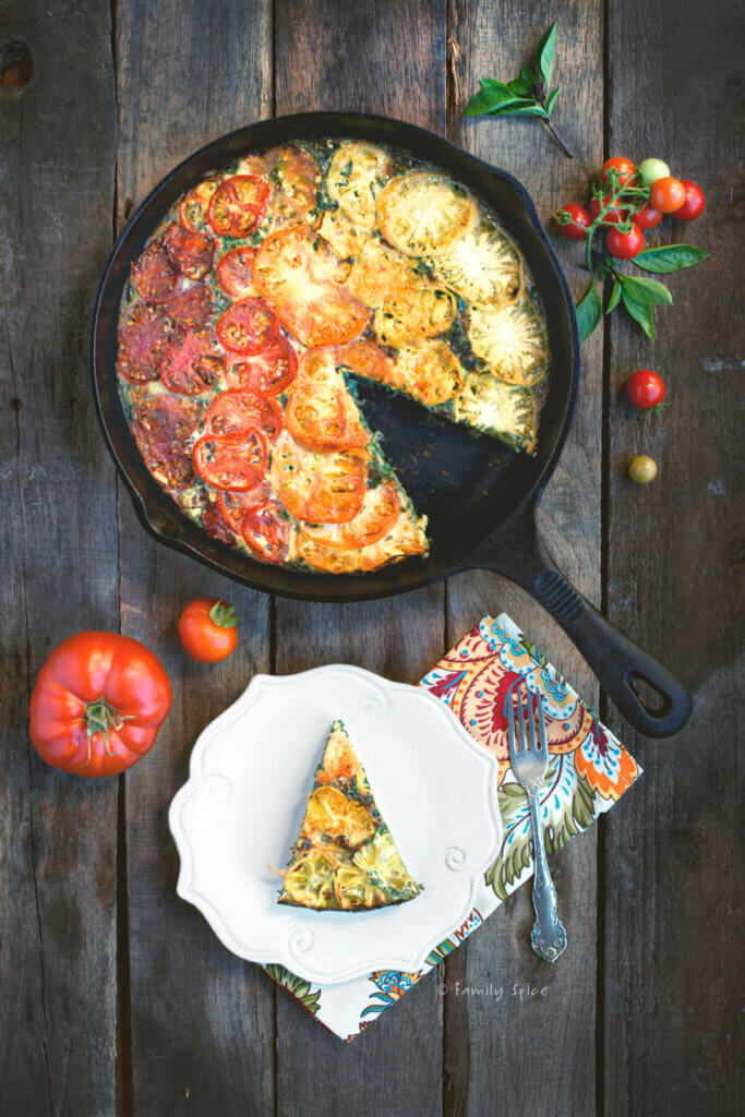 Top view of an heirloom tomato quiche in a cast iron pan with fresh tomatoes and a plate with a slice on it