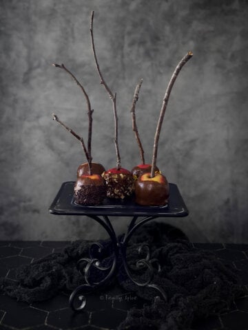 A black metal cake stand with six small caramel apples on it set in a dark and moody setting.