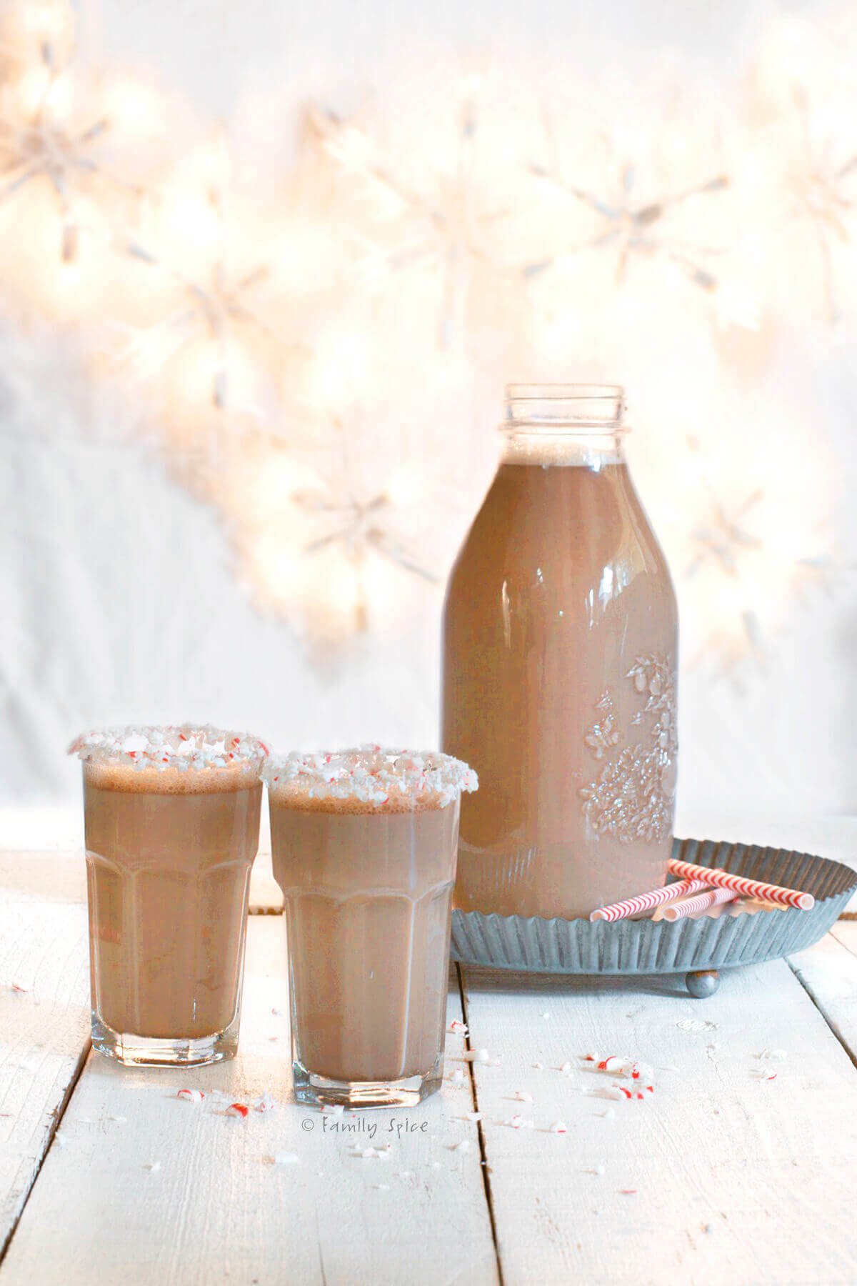 Two glasses of chocolate eggnog with a bottle of it behind it.