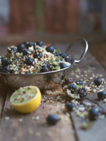 Closeup of a metal bowl with handles with blueberry quinoa salad with feta and lime half next to it