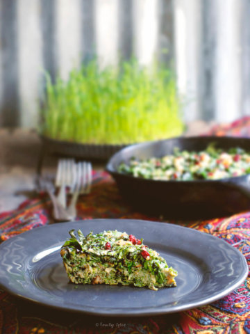 A slice of kuku sabzi (persian herb frittata) on a grey plate with a cast iron pan and a plate of sprouts behind it