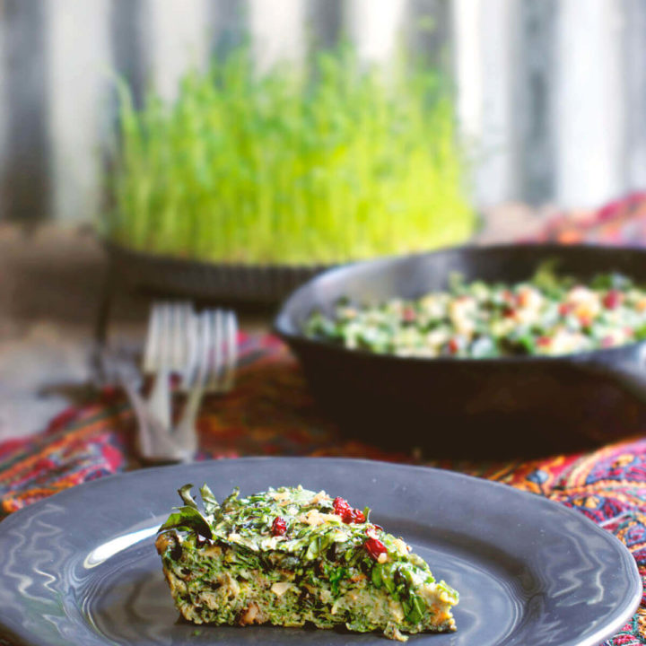 A slice of kuku sabzi (persian herb frittata) on a grey plate with a cast iron pan and a plate of sprouts behind it