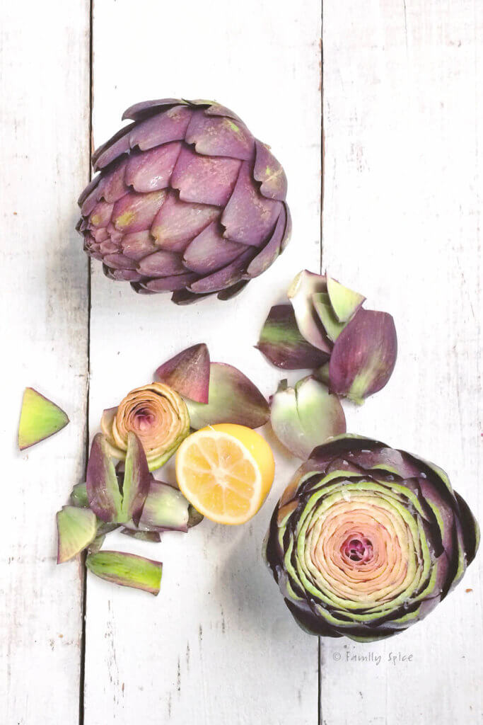 Two purple artichokes, on with the top cut off with some leaves and lemons next to it on a white surface