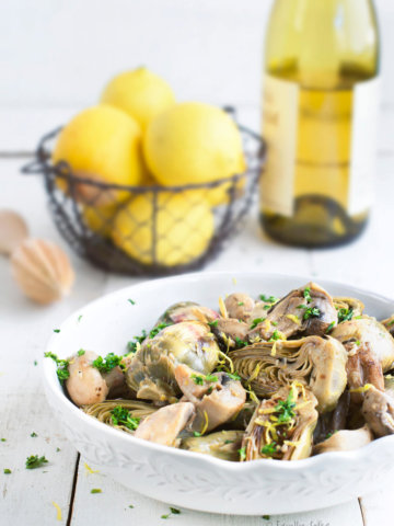 Braised baby artichokes with mushrooms and lemons with a basket of lemons and a bottle of white wine behind it
