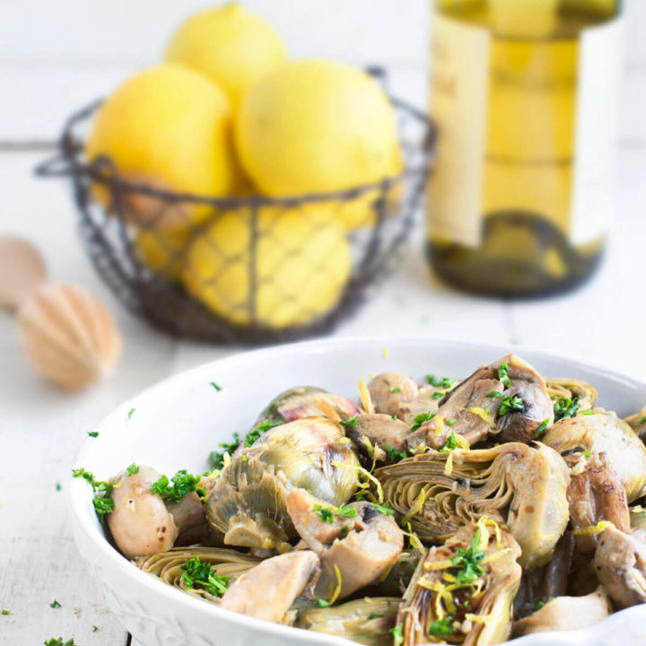 Braised baby artichokes with mushrooms and lemons with a basket of lemons and a bottle of white wine behind it