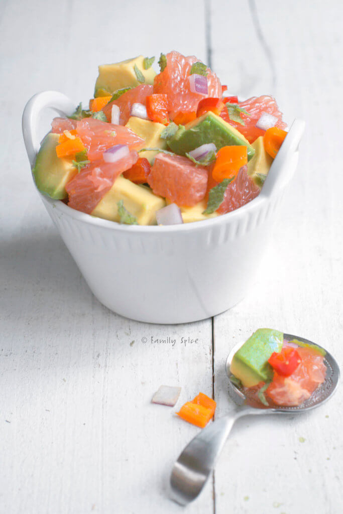 A small white bowl with avocado grapefruit salad with a metal scooping spoon next to it