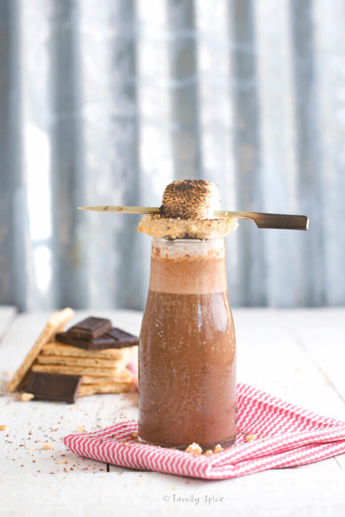 A small glass milk bottle with chocolate s'more smoothie with the rim coated with crushed graham crackers and topped with a marshmallow