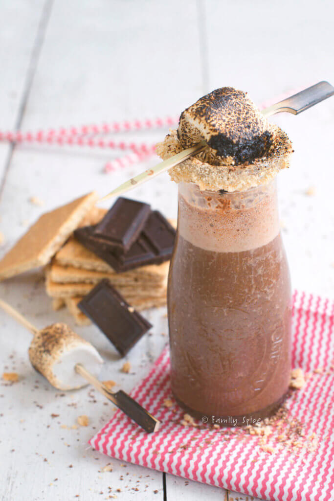 Closeup of a small glass milk bottle with chocolate s'more smoothie with the rim coated with crushed graham crackers and topped with a flaming marshmallow