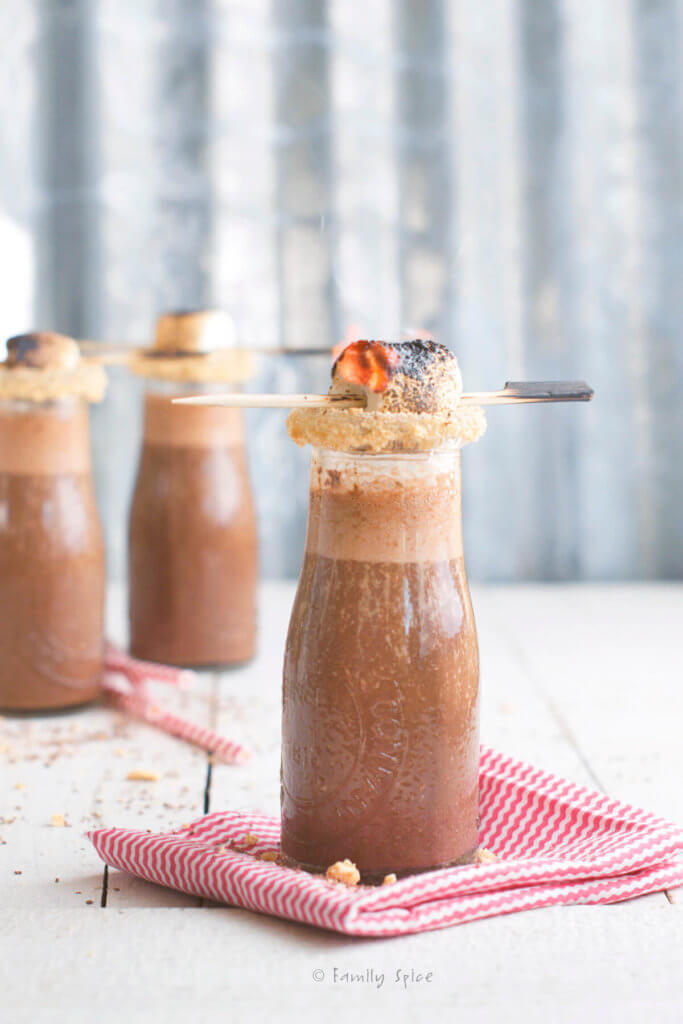 A small glass milk bottle with chocolate s'more smoothie with the rim coated with crushed graham crackers and topped with a flaming marshmallow