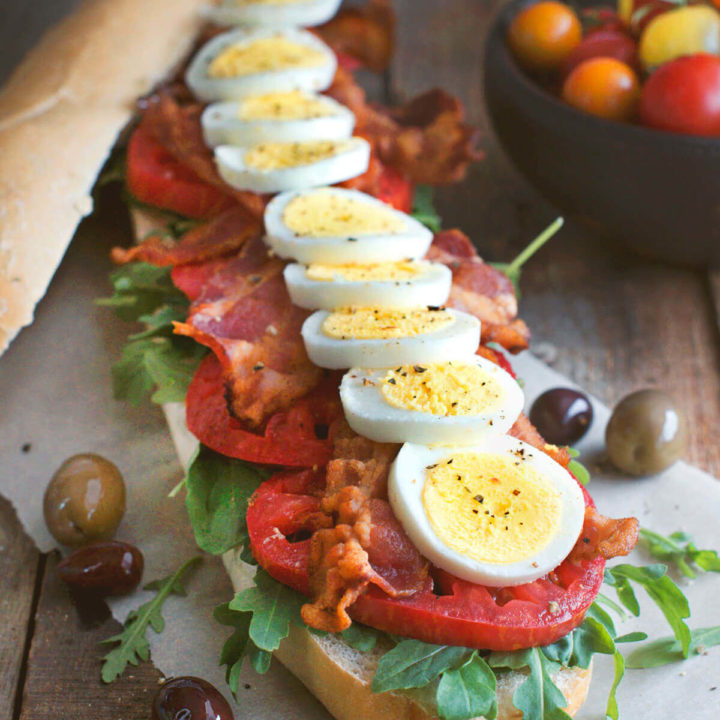 Side view of a baguette filled with slices of hard boiled egg, tomatoes, lettuce and bacon